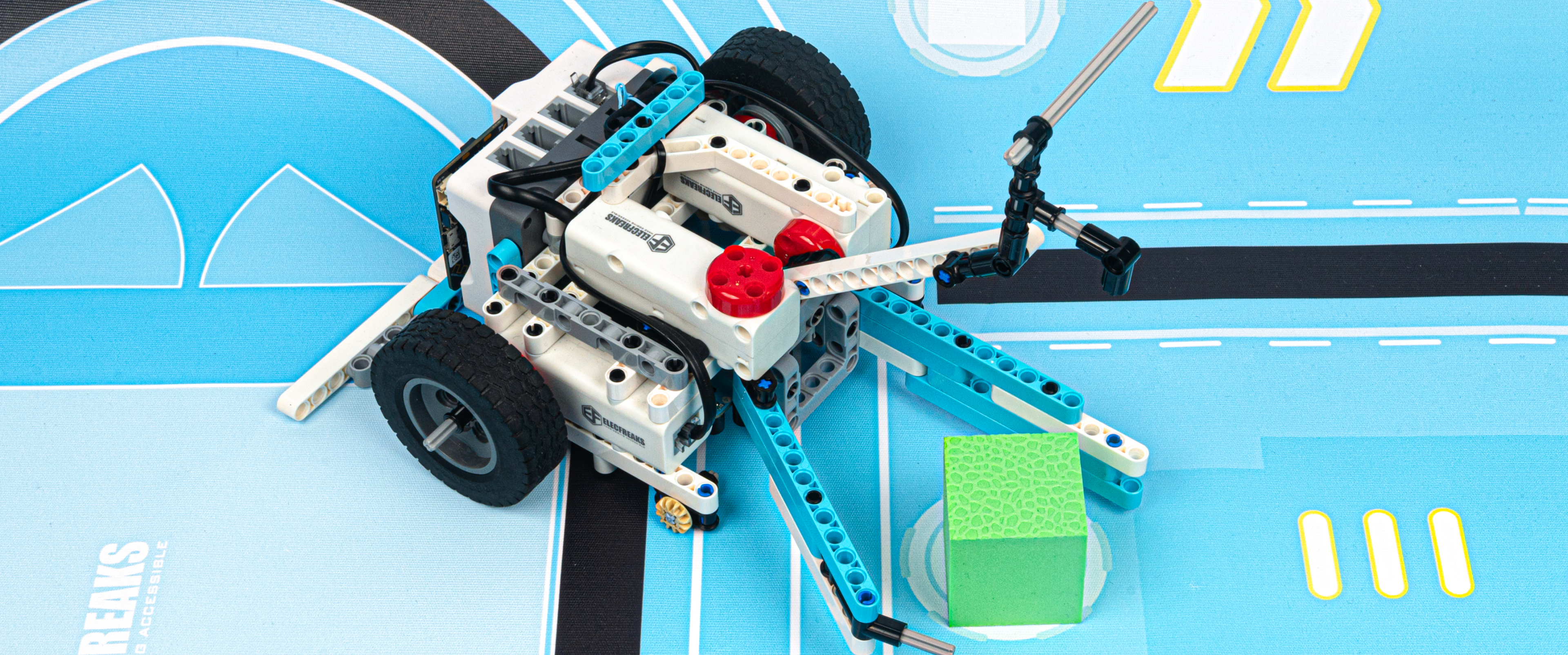 DIY robot kit with a wheeled robot built from building blocks and controllers, performing a task of grabbing a green cube on a competition mat