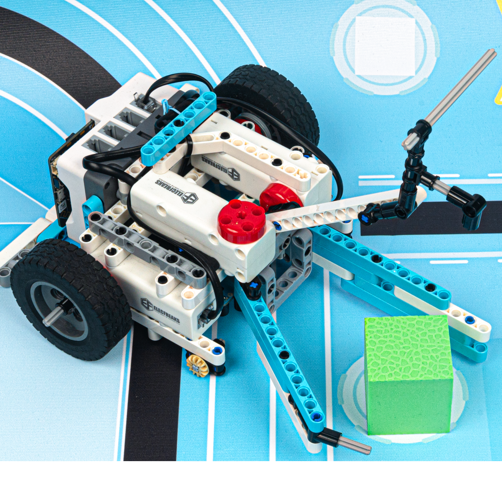 DIY robot kit with a wheeled robot built from building blocks and controllers, performing a task of grabbing a green cube on a competition mat