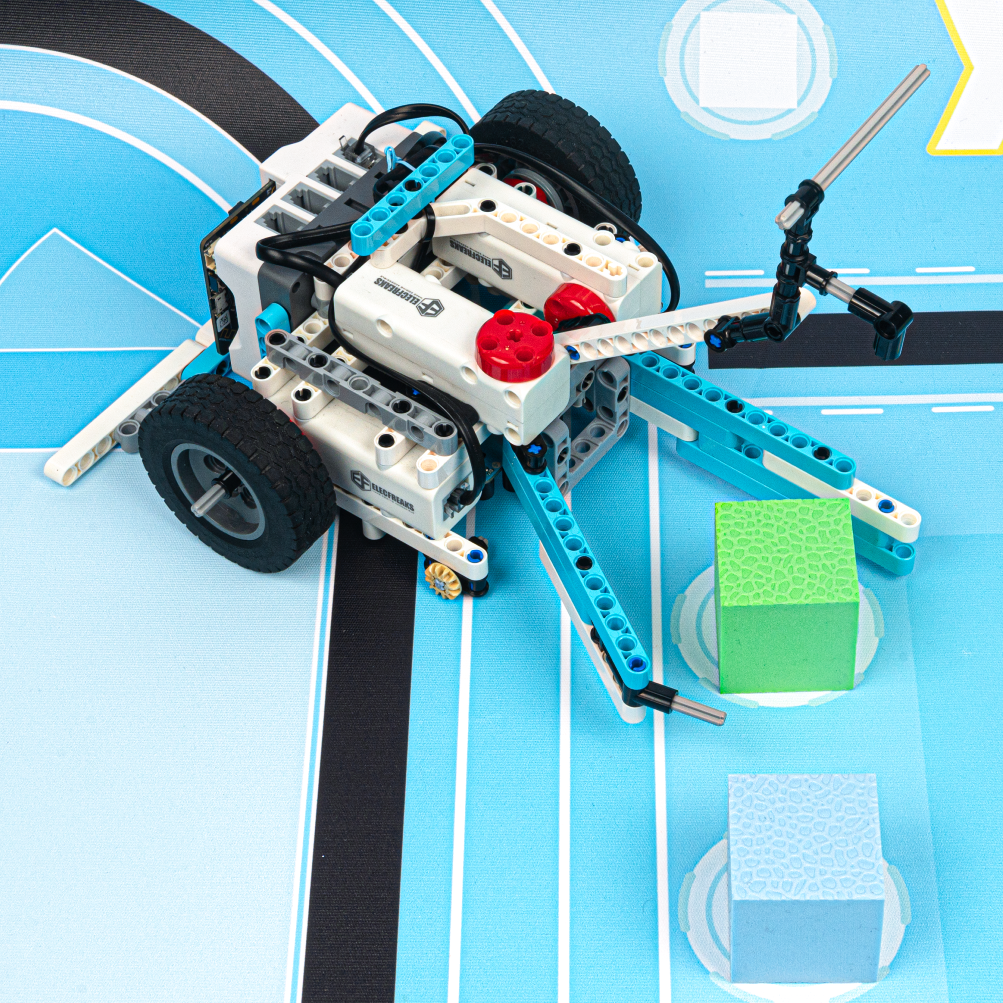 DIY robot kit with a wheeled robot built from building blocks and controllers, performing a task of grabbing a green cube on a competition mat